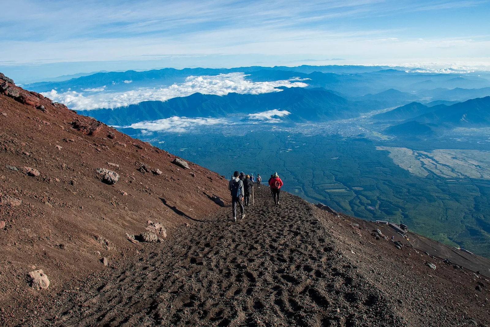 Hikers walking a stone-paved mountain trail through forest toward Mount Fuji